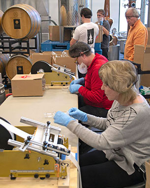 Volunteers wearing gloves assemble and label bottles of hand sanitizer inside the Gervasi Vineyard distillery, surrounded by wine barrels and production equipment.